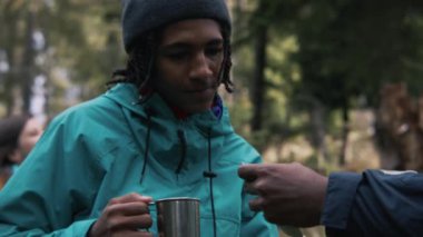 Afrikalı Amerikalı genç yürüyüşçü metal bardağı elinde tutuyor ve konuşuyor. Çeşitli yürüyüş arkadaşları orman kampında dinlenmek için durdular. Doğa keşfi ve turizm kavramı. Aktif boş zaman. Yavaş çekim.