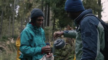 Afrikalı Amerikalı turist metal bardağı elinde tutar, diğer gezgin çaydanlıktan sıcak su döker. Yürüyüş arkadaşları ormanda kamp yapmak için mola verdiler. Doğa keşfi ve turizm. Yavaş çekim.