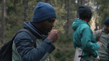 Afro-Amerikalı turist bardağı elinde tutuyor ve sıcak çayı bayan gezginlerle paylaşıyor. Çok ırklı yürüyüş arkadaşları orman kampında dinlenmek için durdular. Doğa keşfi ve turizm. Yavaş çekim.