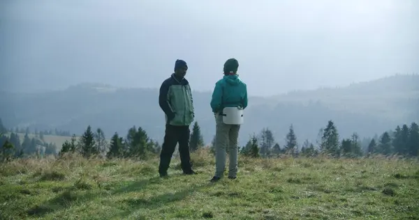 African American tourist and his son stand on top of hill and talk during trek or expedition in the mountains. Family of hikers or travelers rest after long walking trip to forest. Tourism and travel.
