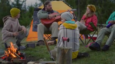 Annesi beyaz bir çocuk kamp ateşinin yanında oturur, marşmelov pişirir ve yürüyüş arkadaşlarıyla konuşur. Bir grup turist ormandaki kampta dinleniyor. Yetişkin Kafkasyalı yürüyüşçü oturur ve gitar çalar. Aktif boş zaman.