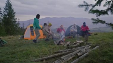 Çeşitli yürüyüşçüler kamp alanında dinlenmek için durdular, ateş yakıp yemek pişirmeye başladılar. Turistler gitar çalar, şarkı söyler ve yemek yerler. Arka planda güneş paneli olan güzel manzara ve çadır.