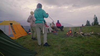 Kampta dinlenen yürüyüş arkadaşlarıyla dolu bir çekim. Afro-Amerikalı adam kamp ateşine lokum pişirmeye geliyor. Kafkasyalı turist gitarla oturur ve arkadaşlarıyla konuşur. Dağlara giden yürüyüşçüler.