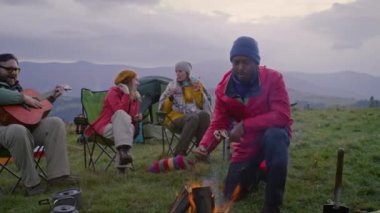 Çok ırklı gezginlerden oluşan bir grup, uzun bir yolculuktan sonra gitar çalıyor, konuşuyor, gülüyor ve yemek yiyor. Açık hava meraklıları kamp alanında dinlenmek ve ateşte yemek pişirmek için durdular. Doğa keşfi ve turizm kavramı.