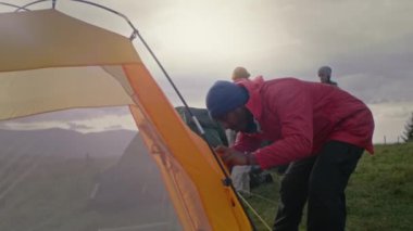 Çok ırklı bir grup açık hava meraklısı kamp kurdu. Afrikalı Amerikalı erkek gezgin çadırı kapatıyor. Turistler uzun bir yolculuktan sonra dinlenmek için durdular. Doğa keşfi ve turizm konsepti. Yavaş çekim.