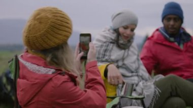 Kafkasyalı kadının oturduğu, telefonuyla fotoğraf çektiği ve arkadaşlarıyla konuştuğu yakın plan. Dişi turist, yürüyüş arkadaşlarıyla selfie çeker. Yürüyüşçüler dağlarda tatil yaparken dinlenirler. Açık hava aktiviteleri.