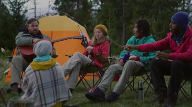 Çeşitli turist grupları kampta marshmallow pişirir ve konuşur. Yetişkin beyaz adam oturur ve gitar çalar. Yürüyüş arkadaşları güzel ormandaki kamp alanında dinleniyor. Yürüyüşçüler dağlara giderken.