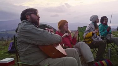 Çok ırklı gezginlerden oluşan bir grup gitar çalıyor, birlikte konuşuyor ve uzun bir yolculuktan sonra yemek yiyor. Turistler kamp alanında dinlenmek ve ateşte yemek pişirmek için durdular. Açık hava keşfi ve turizmi kavramı.