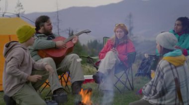 Çeşitli yürüyüşçüler kamp ateşinin yanındaki kampta dinleniyor. Yürüyüş arkadaşları konuşur, yemek yapar, lokum yer ve müzik dinler. Yetişkin beyaz adam oturur ve gitar çalar. Dağlardaki kamp alanında bir grup turist.