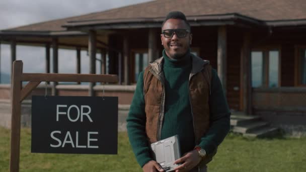 拥有数字平板电脑的非洲裔美国地产经纪人站在旁边等待销售标志 看着相机 微笑着 真正的国家工作人员在等现代住宅的来访者 住宅物业出售概念 — 图库视频影像