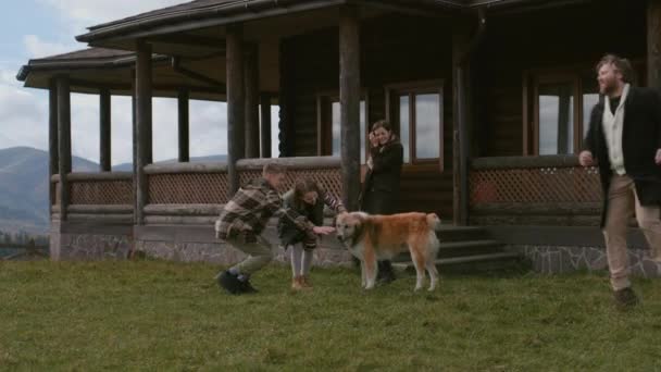 一对带着孩子的成年夫妇走在屋前 和狗在草坪上玩耍 幸福的家庭定居在山里租来的现代小木屋里度假 或搬到新的住宅 房地产的概念 — 图库视频影像