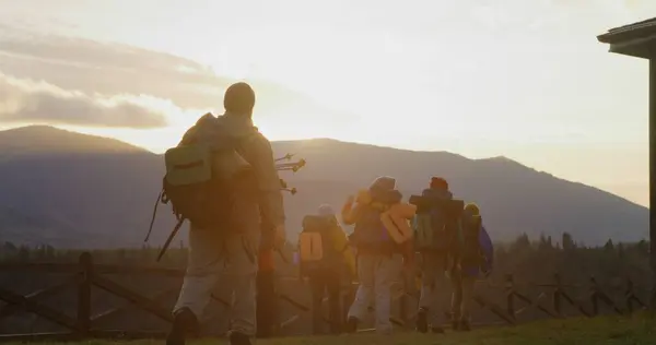 Diverse hikers with backpacks and trekking poles go hiking in mountains. Happy tourist family or hiking buddies during vacation road trip. Sunset and mountain landscapes in background. Slow motion.