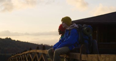 Ahşap çitlerin yanındaki çok ırklı genç turistler, gün batımını ve doğanın tadını çıkaran dağ manzaralarını seyrediyorlar. Yürüyüşten ya da dağlarda keşfe çıktıktan sonra dinlenen iki genç çocuk. Yavaş çekim.