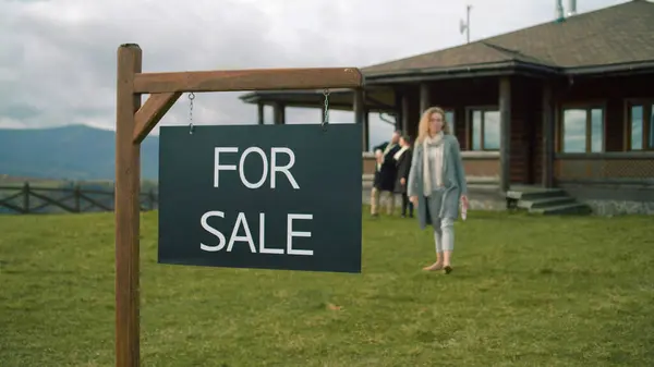 For sale sign on beautiful green lawn. Female real estate agent comes and puts sold sticker on sign. Stylish house with traditional architecture in mountains. Residential property on sale concept.
