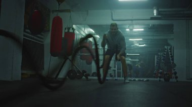 Sporcu adam karanlık boks salonunda halatlarla egzersiz yapıyor. Profesyonel erkek boksör şampiyonluk maçından önce kardiyo ya da dayanıklılık egzersizi yapar. Fiziksel aktivite ve yoğun CrossFit eğitimi.
