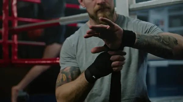 Adult man sits near boxing ring and wraps his hands with bandage for martial arts. Athlete prepares to fight, competition or training in box gym. Boxer performs shadow boxing at background. Close up.
