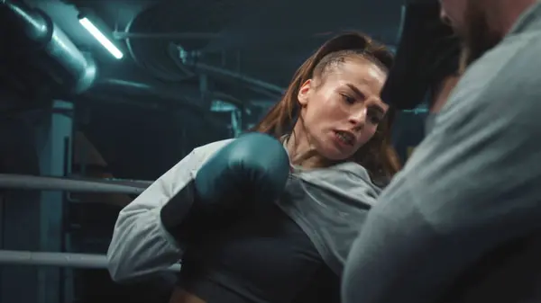 Close up of female fighter in boxing gloves practicing fighting technique and exercising before match in gym. Athletic boxer hits punching mitts on boxing ring. Physical activity and workout concept.