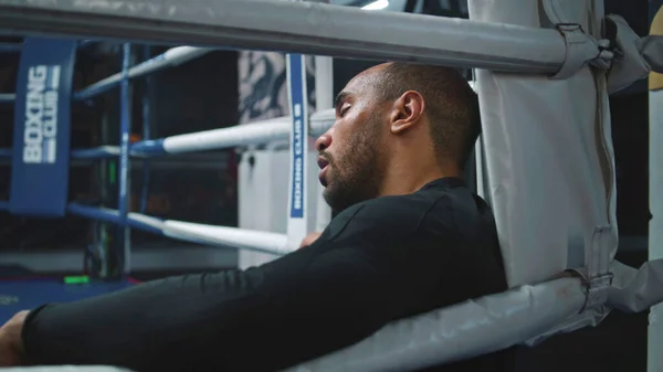 Tired and exhausted African American boxer sits in boxing ring corner. Fighter with beads of sweat on face rests after training. Athlete prepares to tournament or competition in boxing gym. Close up.