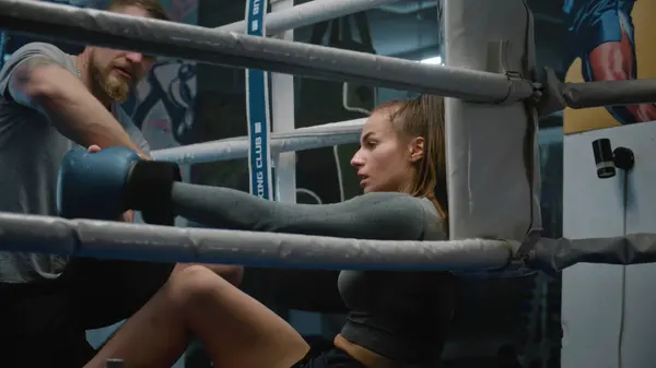 Tired boxer sits at ring corner after long fight. Male trainer comes to woman and helps her to take off boxing gloves and bandages. Athlete prepares to fight, competition or training in boxing gym.