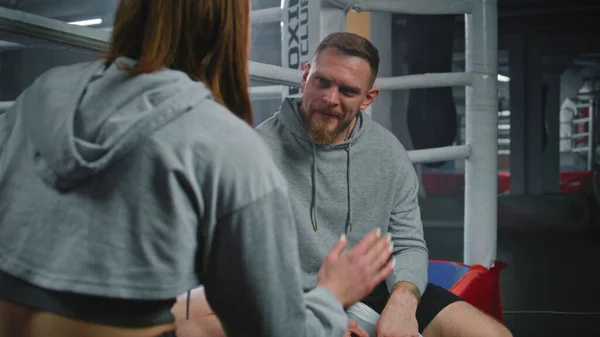 Female boxer sits on ring and talks with male trainer about fight. Coach consults boxer and explains fighting technique. Woman prepares to fighting competition in gym. Physical activity and workout.