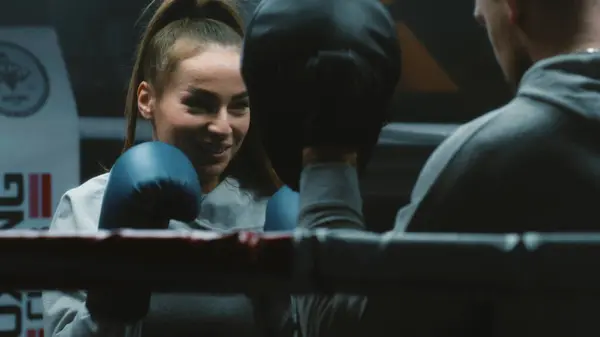 Close up of female boxer in boxing gloves exercising with male coach and preparing to fighting match in dark gym. Athletic woman hits punching mitts on boxing ring. Physical activity and workout.