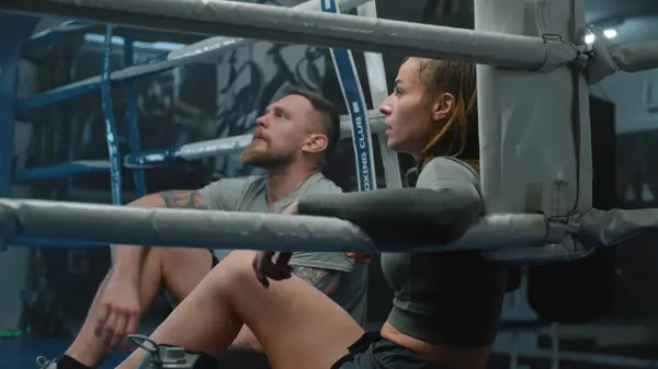 Tired woman sits at ring corner, takes off boxing gloves and starts drink water. Male trainer sits down near boxer and discusses training. Athlete prepares to fight or competition in boxing gym.