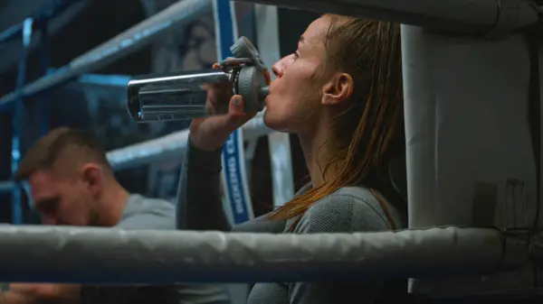 Tired woman sits at ring corner, takes off boxing gloves and starts drink water. Male trainer sits down near boxer after hard train. Female athlete prepares to fight or competition in boxing gym.