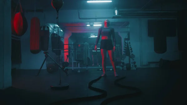 Professional female boxer does cardio or endurance workout before championship fight. Strong woman exercises with ropes in dark boxing gym with LED lighting. Physical activity and CrossFit training.