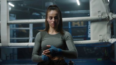 Yoğunlaşmış kadın boks ringinin yanında duruyor ve ellerini dövüş sanatları için bandajla sarıyor. Sporcu boks salonunda dövüşmeye, yarışmaya ya da antrenmana hazırlanır. Erkek boksör arka planda ısınıyor.