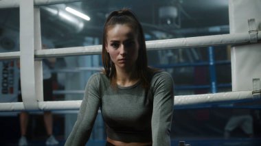 Kadın boks ringinin yanında oturuyor, ellerini sargıyla sarmalayıp kameraya bakıyor. Sporcu boks salonunda dövüşmeye, yarışmaya ya da antrenmana hazırlanır. Boksörler arka planda ısınıyor. Portre.