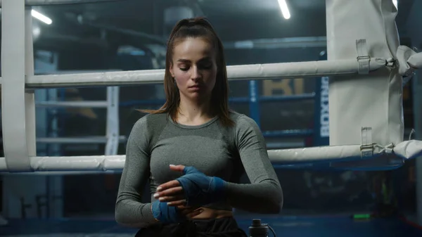 Concentrated woman stands near boxing ring and wraps her hands with bandage for martial arts. Athlete prepares to fight, competition or training in box gym. Male boxer performs warm up at background.