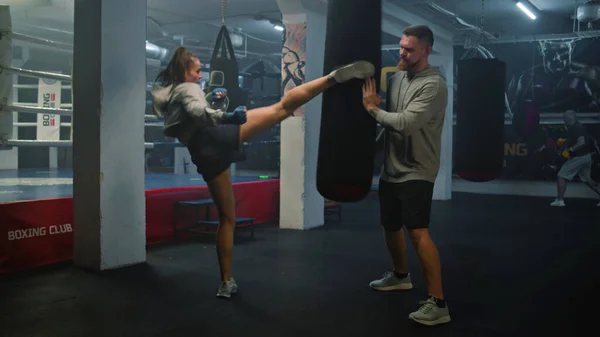 Female kickboxer in boxing bandages hits punching bag while training in dark boxing gym. Athletic woman practices and prepares for match with male coach. Physical activity and intensive workout.
