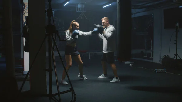 Female boxer in boxing gloves hits punching mitts and practices fighting techniques with coach in dark boxing gym. Athletic woman exercises and prepares for match. Physical activity and workout.