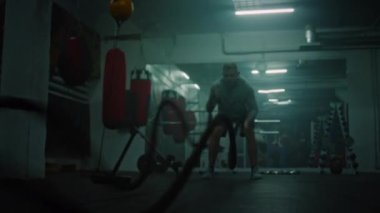 Sporcu adam karanlık boks salonunda halatlarla egzersiz yapıyor. Profesyonel erkek boksör şampiyonluk maçından önce kardiyo ya da dayanıklılık egzersizi yapar. Fiziksel aktivite ve yoğun CrossFit eğitimi.