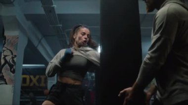 Boks bandajlı kadın boksör maçtan önce erkek antrenörle spor yapıyor. Atletik kadın kum torbasına vuruyor ve dövüş tekniği uyguluyor. Fiziksel aktivite ve egzersiz