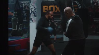 Kendine güvenen Afrikalı Amerikalı boks salonunda antrenman yaparken kum torbasına vurur. Boks eldivenli sporcu turnuvadan önce koçla egzersiz yapıyor. Fiziksel aktivite ve yoğun çalışma.