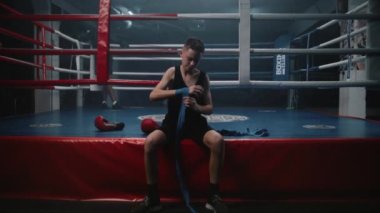 Genç boksör boks ringinin yanında oturur ve ellerini dövüş sanatları için bandajla sarar. Genç sporcu turnuvaya ya da yarışmaya hazırlanıyor, boks salonunda antrenman yapıyor. Fiziksel aktivite. Dolly vurdu..