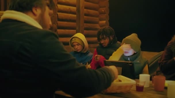 一群多种族的旅行者晚上坐在木制的凉亭里 边吃饭边聊天 小女孩用平板电脑 度假期间 游客朋友们在度假别墅外放松一下 — 图库视频影像