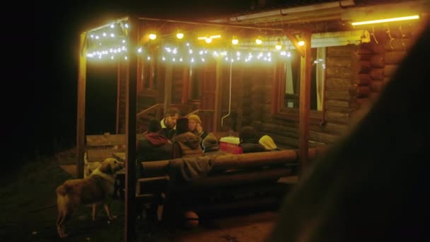 観光客のグループは夕方に木製のガゼボに座り 夕食を食べながら話をする 多民族の友人や旅行者の家族 休日の家の外で週末の田舎で犬と一緒に — ストック動画