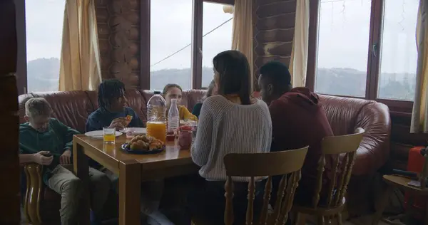 Multicultural family sit around table, eat, drink, talk and clink glasses. Friends gathered together in holiday home to rest. Tourists relax in wooden cottage during their vacation. Active leisure.