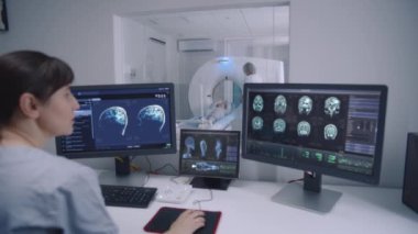 Kadın radyolog kontrol odasında oturur ve beyin taraması sonuçlarıyla monitörlere bakar. Profesyonel doktor MRI, CT ya da PET taramalarını kontrol ederken arka planda hasta işlem görüyor.