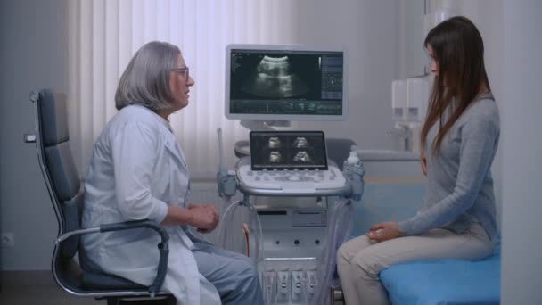白人女性は病院のソノグラフィー室に座り 医師に腹痛を訴えています 医師は検査前に女性患者と話をする 背景のモニターが付いている超音波機械 — ストック動画
