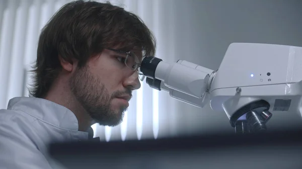 Medical worker looks under microscope, studies and analyzes bacteria or blood sample in modern laboratory. Male scientist works in advanced lab, does biotechnology or pharmaceutical research. Portrait