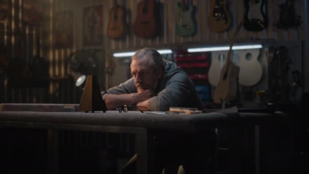 疲れた職人 職人は 楽器を作るためのスタイリッシュなワークショップに座っているメトロノームを見ます 成熟した大工は長い勤務日の後に休む 木製のギターが壁に掛かっている ハンドメイドコンセプト — ストック動画