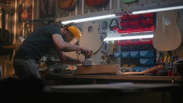 保護フェイスシールドの男性カーペンターは 研磨機を使用して木製ギターボディを磨きます ウッドワーキングマスター 職人は現代のワークショップで楽器を作ります ハンドメイドと起業家精神 — ストック動画