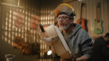 Güvenli yüz kalkanındaki olgun marangoz cilalamadan önce gitar gövdesinden ahşabı temizler. Modern atölyede profesyonel bir zanaatkar müzik aleti yapıyor. İş arkadaşı arka planda çalışır.