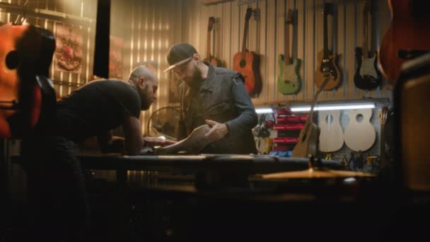 2人のプロの大工が木製ギターボディを空白で点検し 現代のワークショップで楽器を作るプロセスを議論します コリーグはバックグラウンドで活動しています ハンドメイドと起業家のコンセプト — ストック動画
