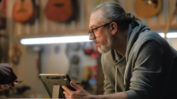 用数字平板电脑将成熟的男性木匠近距离地绘制出吉他的蓝图 纹身工匠与同事交谈 并用口径小巧时尚作坊测量吉他手的身体 — 图库视频影像