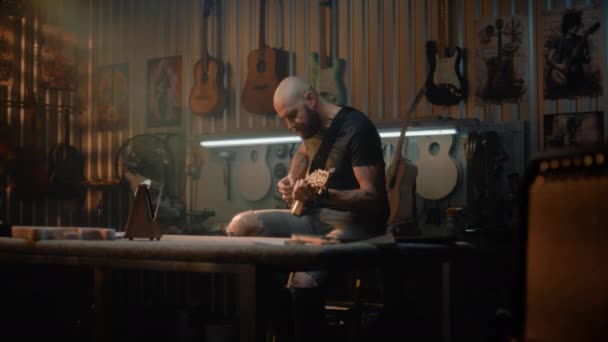 男性ミュージシャンは 楽器を作るためのワークショップでメトロノームとギターを演奏します プロの大工や職人が手作りの製品をチェックします 木製ギターは背景の壁に掛かります — ストック動画