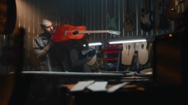 Erkek zanaatkar akustik gitar çalar, sesi kontrol eder. Profesyonel zanaatkar modern atölyede ahşaptan müzik aleti yapar. İş arkadaşı arka planda çalışıyor. El işi ve küçük işletmeler..